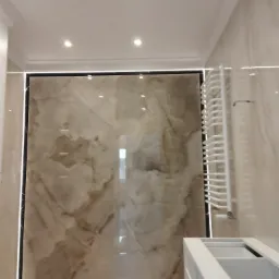 Wykończona łazienka z beżowymi płytami imitującymi marmur, podświetlanymi listwami LED i białym grzejnikiem drabinkowym, widoczna częściowo zabudowa umywalki.