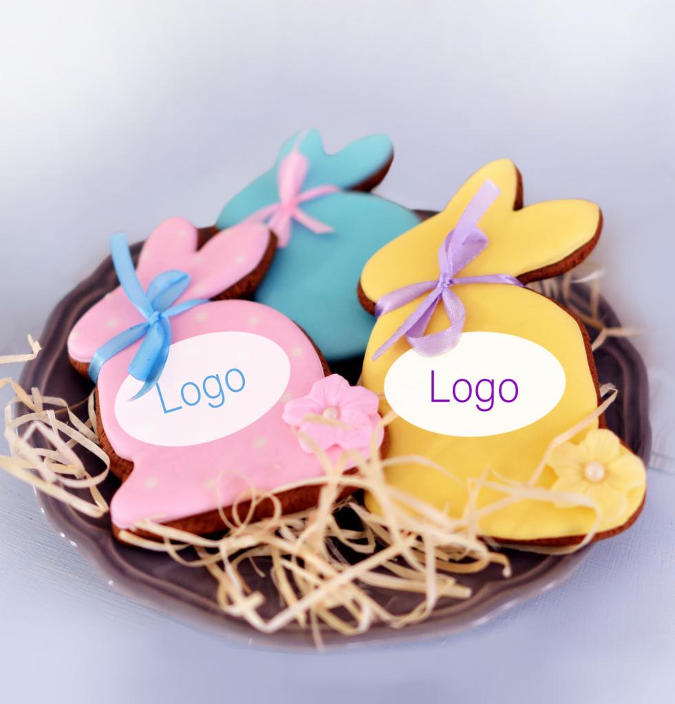 Ręcznie robione, kolorowe ciasteczka w kształcie zajączków wielkanocnych z lukrem i wstążkami, ułożone na szarym talerzu z dekoracyjnym siankiem, z naniesionym napisem 'Logo' na każdym ciasteczku.