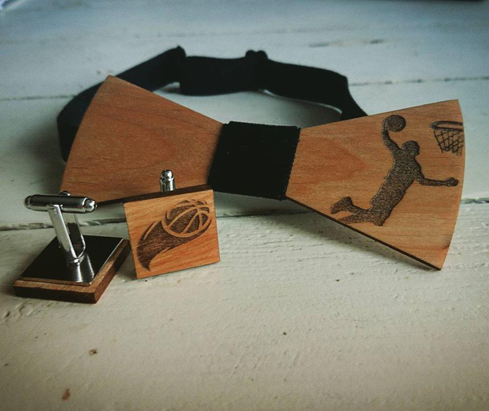 Drewniana mucha i spinki do mankietów z motywem koszykarza i piłki, leżące na białym, drewnianym stole.