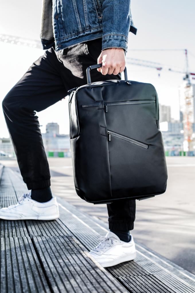 Mężczyzna w dżinsowej kurtce i białych sneakersach wchodzi po drewnianych schodach, trzymając czarny, minimalistyczny plecak-walizkę na kółkach z wysuwaną rączką, na tle miejskiego krajobrazu...