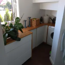 Mała pralnia z białą pralką, drewnianym blatem, białymi szafkami, koszem wiklinowym, żelazkiem i rośliną w doniczce przy oknie.