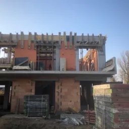 Surowy stan budowy domu z czerwonej cegły, widoczne szalunki na stropie i nadprożach, stosy płyt szalunkowych na pierwszym planie, błękitne niebo w tle.