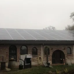 Instalacja paneli słonecznych na dachu budynku z cegły, widoczny fragment rusztowania i elementy wyposażenia ogrodu. Mglista pogoda.