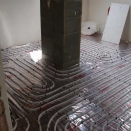 Rozłożone rury grzewcze na podłodze z siatką montażową, przygotowane do zalania betonem, widoczny fragment kolumny konstrukcyjnej i płyta z widoczną strukturą.