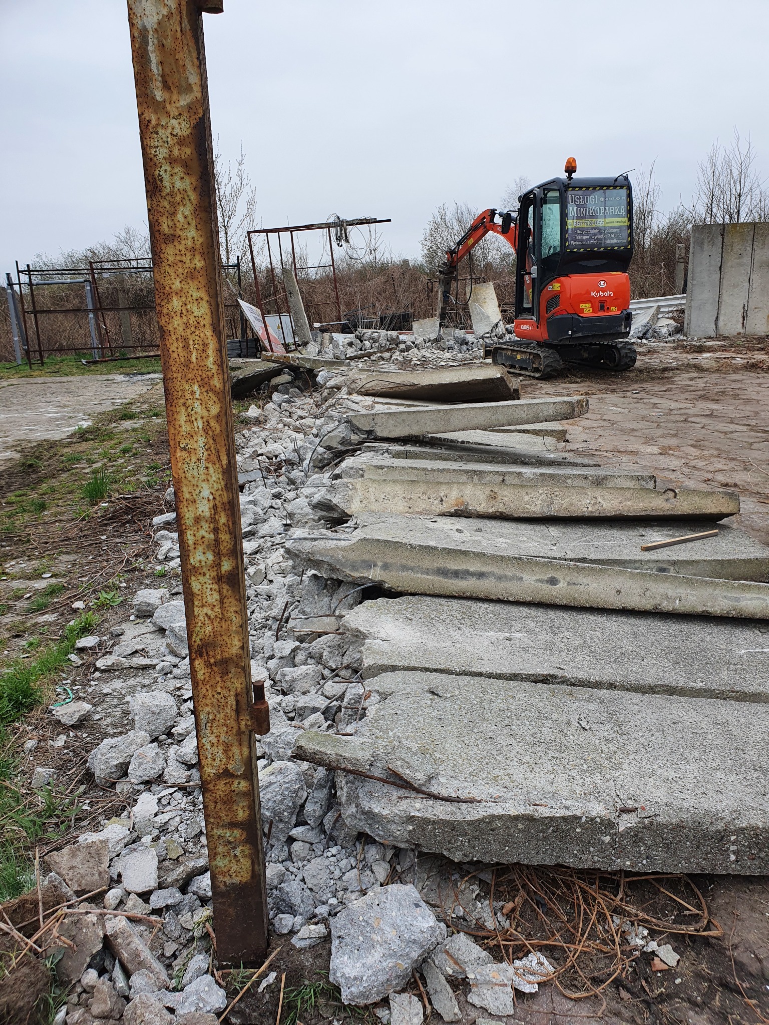 Pomarańczowa minikoparka Kubota kruszy betonowe płyty na gruzowisku, w tle zardzewiały słup i metalowe ogrodzenie.