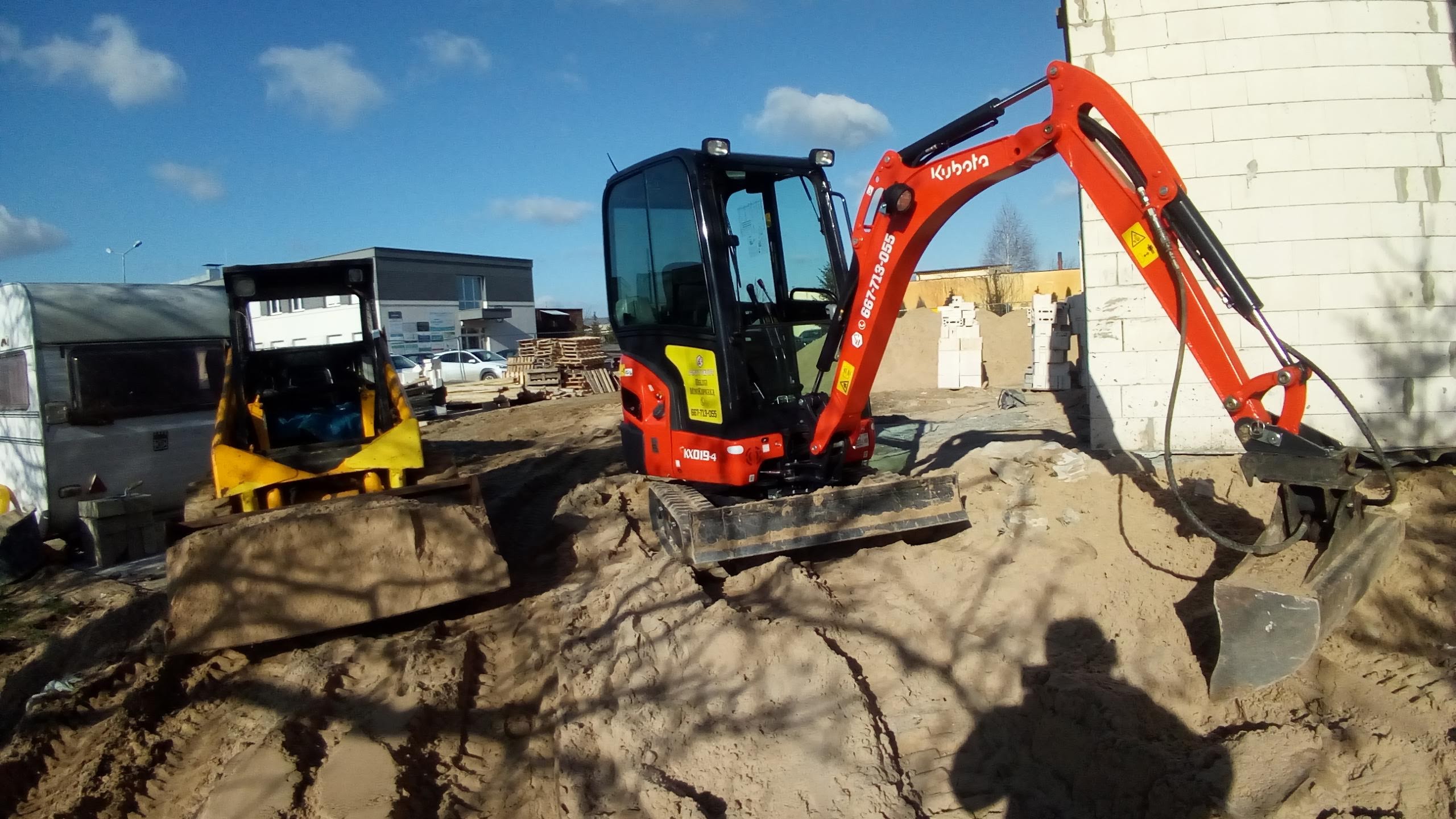 Pomarańczowa mini koparka Kubota na piaszczystym terenie budowy, obok żółta ładowarka, w tle budynki i błękitne niebo z chmurami.