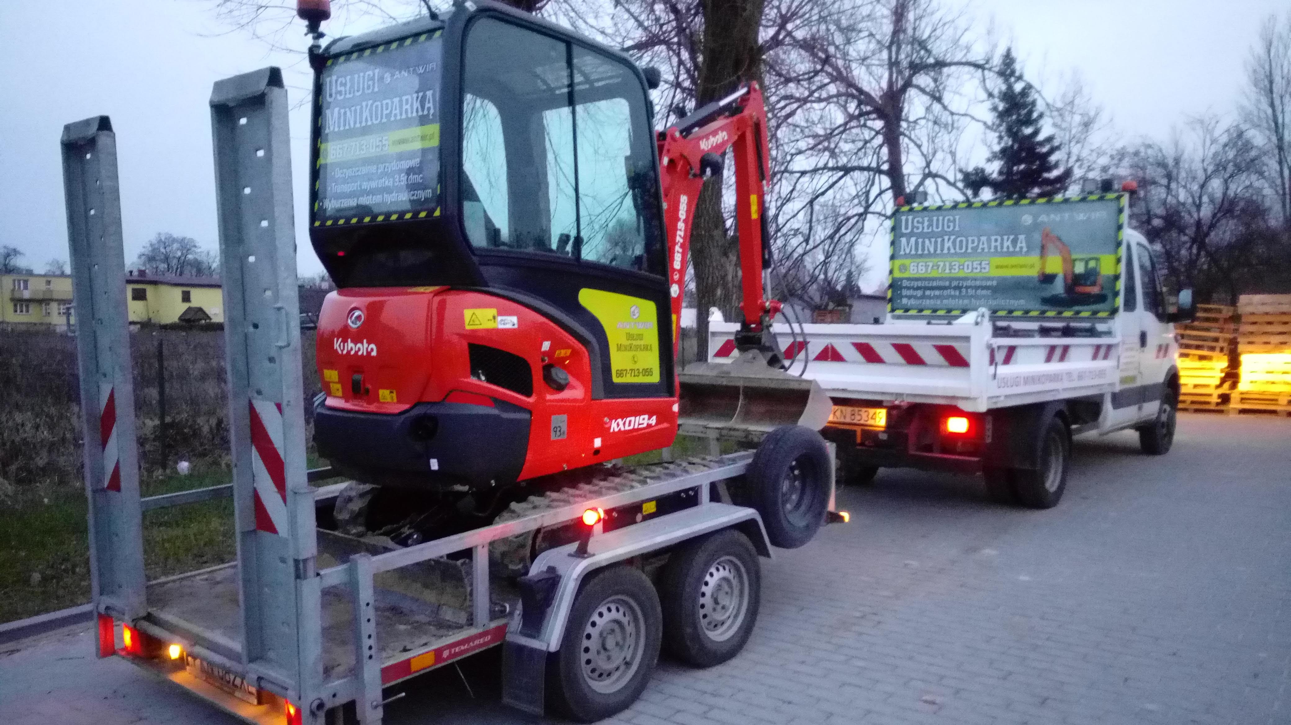 Czerwona mini koparka Kubota KX019-4 na niskopodwoziowej przyczepie transportowej Temared, zaparkowana za białą wywrotką z reklamą usług minikoparką i numerem telefonu, jesienna szaruga.