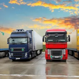 Cztery ciężarówki różnych kolorów (biała, niebieska, czerwona i żółta) zaparkowane w rzędzie na betonowym placu z widocznym odbiciem w kałużach po deszczu, na tle zachodzącego słońca z chmurami.