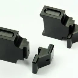 Trzy czarne, precyzyjnie obrobione elementy z metalu lub tworzywa sztucznego na białym tle. Widoczne ślady obróbki skrawaniem, otwory i zagłębienia. Jeden z elementów ma charakterystyczne podłużne...