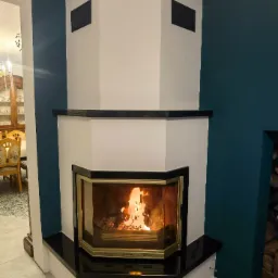 Narożny wkład kominkowy z czarnym obramowaniem i płomieniami, na tle ściany w kolorze butelkowej zieleni i fragmentu jadalni.