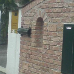 Murowana z cegły elewacja budynku z zaokrąglonym dachem, wnęką w kształcie łuku, skrzynką pocztową i tabliczką informacyjną.