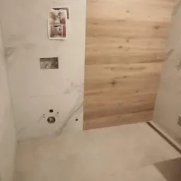 Niedokończona łazienka z białymi marmurkowymi płytkami i drewnopodobnymi panelami ściennymi, widoczne otwory na armaturę i wystające przewody elektryczne.