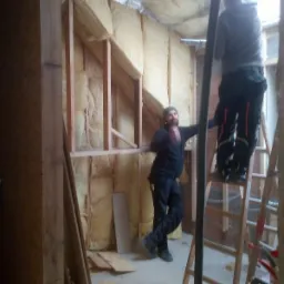 Dwaj mężczyźni pracują nad ociepleniem poddasza wełną mineralną, widoczne drewniane ramy konstrukcyjne i folia aluminiowa na suficie. Jeden z nich stoi na drabinie, drugi opiera się o ścianę.