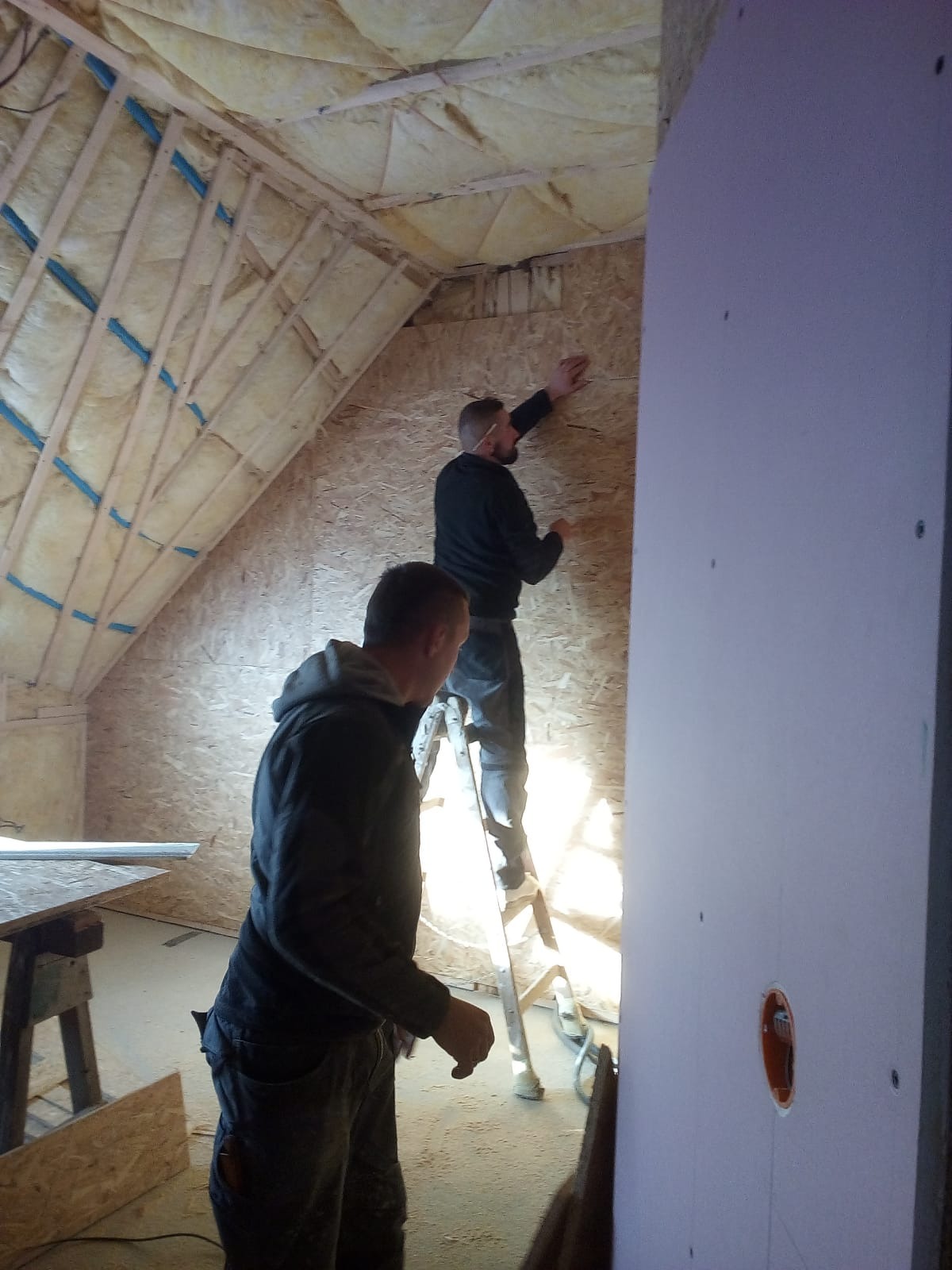 Dwóch mężczyzn na drabinie montuje płyty gipsowo-kartonowe na poddaszu z ociepleniem, widoczna instalacja elektryczna.