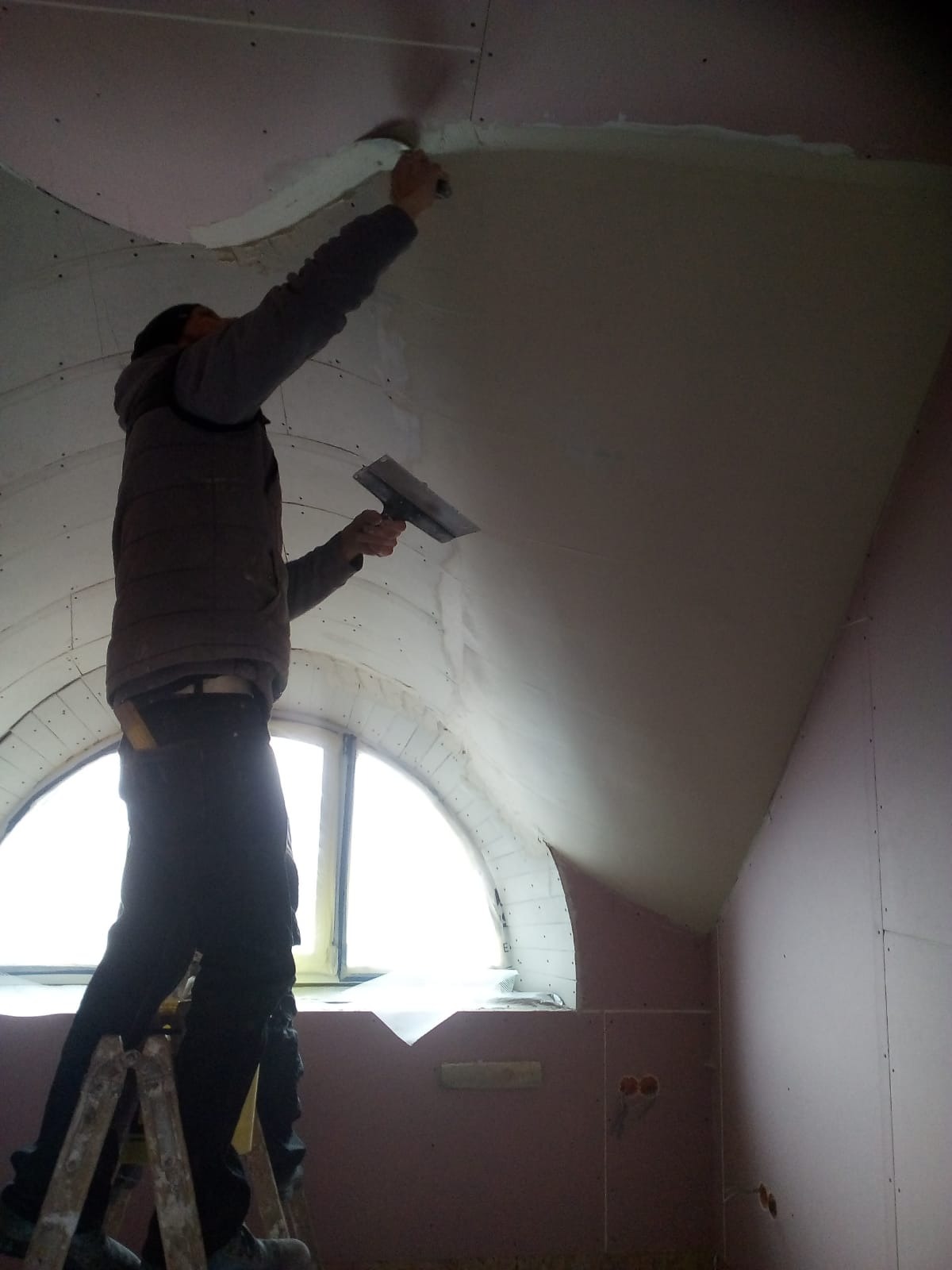 Pracownik na drabinie szpachluje łączenie płyt karton-gips na suficie poddasza z oknem łukowym, widoczne narzędzia i nierówności na powierzchni.