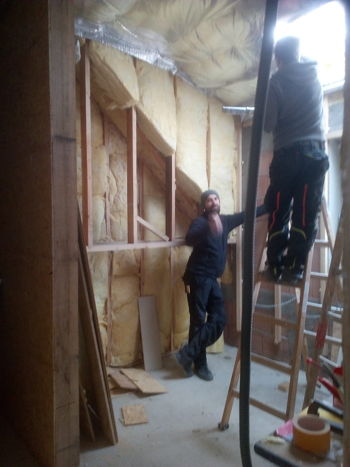 Dwaj mężczyźni pracują nad ociepleniem poddasza wełną mineralną, widoczne drewniane ramy konstrukcyjne i folia aluminiowa na suficie. Jeden z nich stoi na drabinie, drugi opiera się o ścianę.