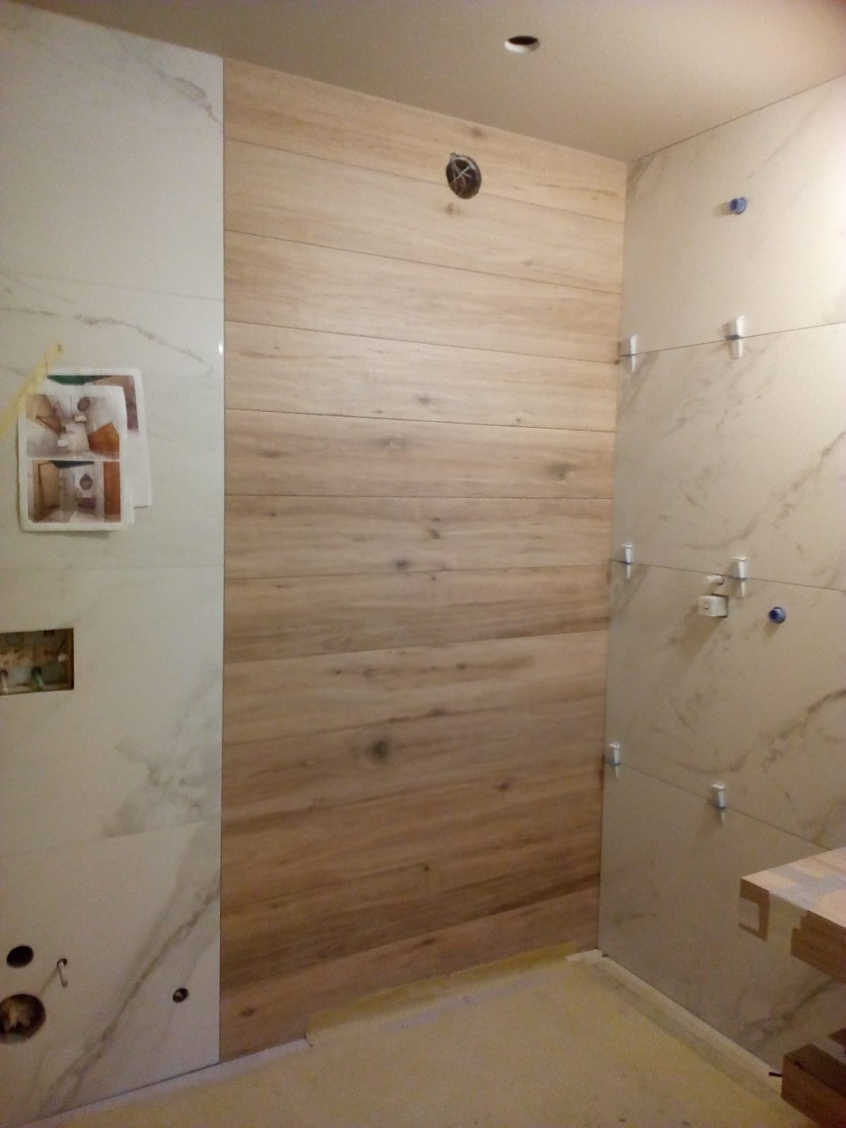 Ściana łazienki wyłożona płytkami imitującymi drewno, sąsiadująca z płytkami marmuropodobnymi, widoczne krzyżyki dystansowe i przygotowane otwory na armaturę.