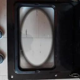 Otwarta, brudna szyba piekarnika z czarną ramą, widoczna podłoga z szarych płytek i fragment buta.