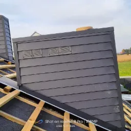 Montaż obróbki blacharskiej komina z szarej blachy na dachu z drewnianą więźbą i papą, widoczny krajobraz polny w tle.