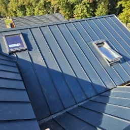 Nowoczesny, grafitowy dach panelowy z dwoma oknami dachowymi, widok z góry na tle zieleni.