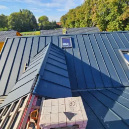 Nowe, grafitowe krycie dachu z blachy na rąbek stojący, z oknami dachowymi, widok z góry na częściowo ukończony dach, z widoczną więźbą i materiałami budowlanymi.