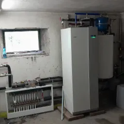 Biała jednostka wewnętrzna z podłączonymi rurami, zbiornik wyrównawczy, rozdzielacz ogrzewania podłogowego w piwnicy z oknem i widocznymi śladami wilgoci na ścianach.