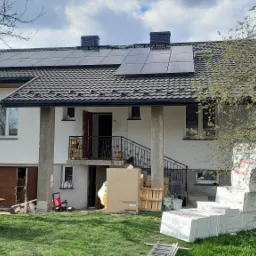 Dwupiętrowy dom z zainstalowanymi panelami słonecznymi na dachu, widoczne materiały budowlane i narzędzia na trawniku, wskazujące na trwające prace instalacyjne.