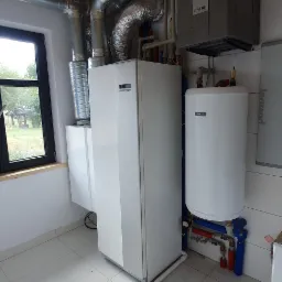 Biała jednostka centralna z szarym modułem wentylacyjnym NIBE, rurami i zbiornikiem na wodę w pomieszczeniu z drewnianym sufitem i oknem z widokiem na zewnątrz.