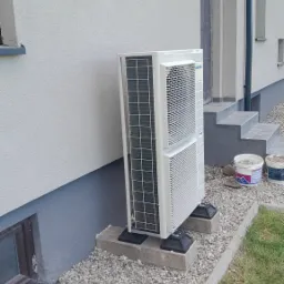 Zewnętrzna jednostka klimatyzacyjna marki Panasonic zamontowana na betonowych podstawach przy ścianie budynku z oknem piwnicznym i schodami wejściowymi, widoczne dwa wiadra z farbą.