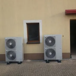 Dwa szare urządzenia grzewcze z podwójnymi wentylatorami, zamontowane na zewnątrz budynku z oknami, na brukowanej powierzchni. W tle widoczne drewniane elementy konstrukcyjne.