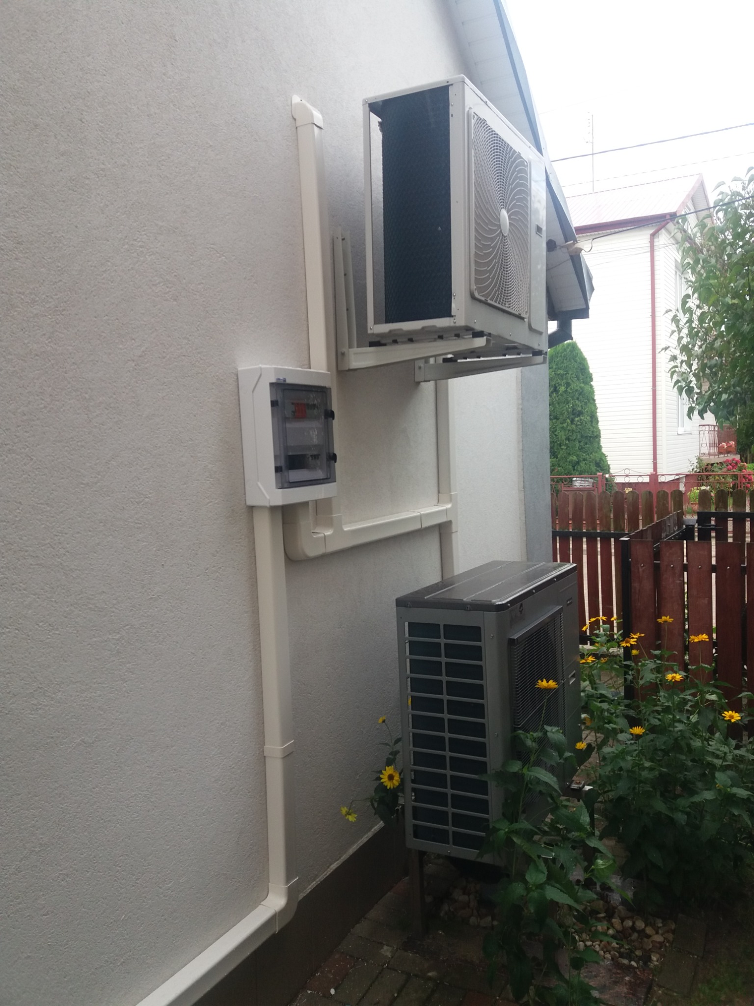 Dwa moduły klimatyzacji zamontowane na ścianie budynku, połączone białymi rurami maskującymi, z widoczną skrzynką elektryczną i żółtymi kwiatami w tle.