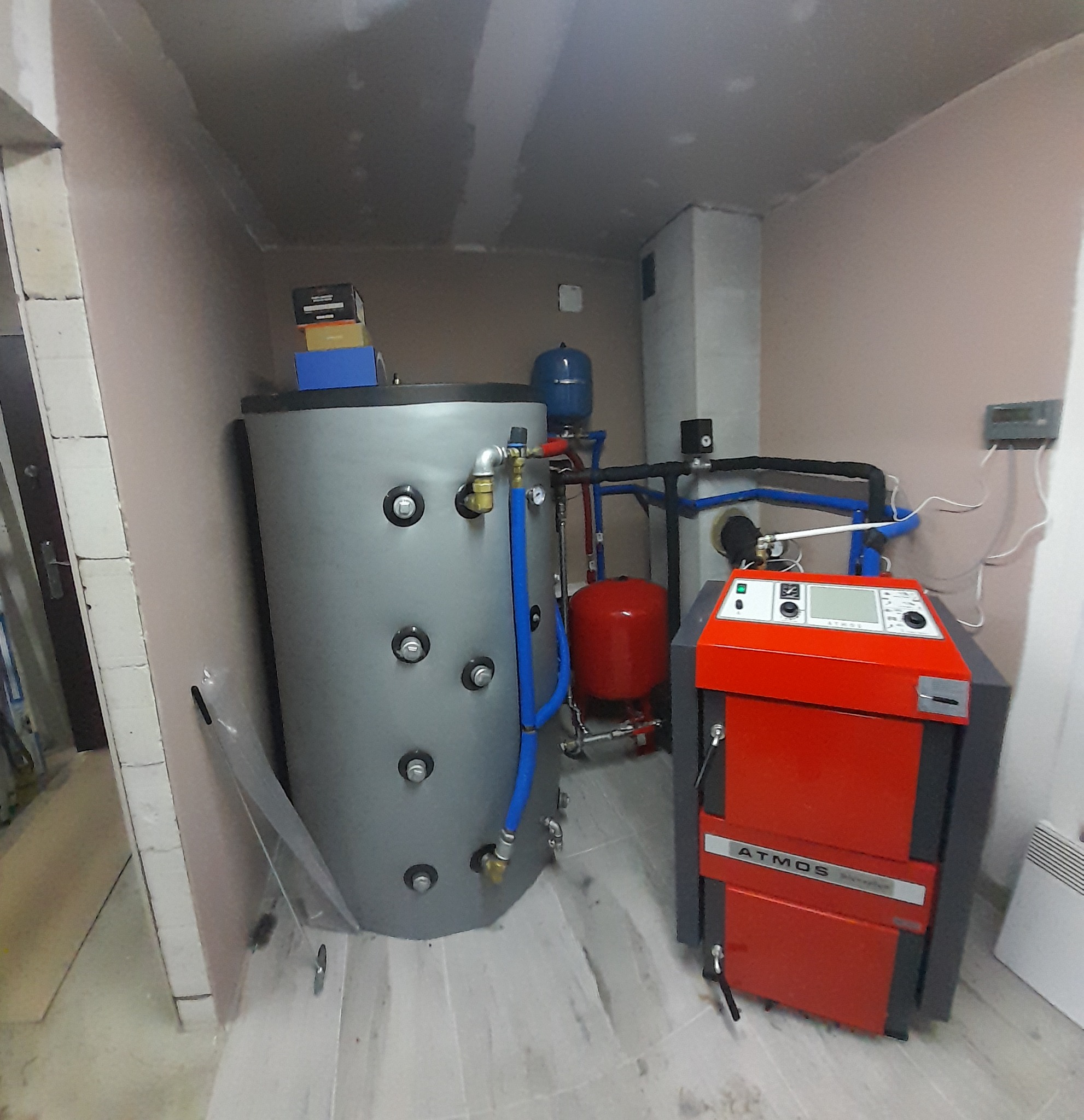 Szary akumulator ciepła z podłączeniami hydraulicznymi i czerwony piec Atmos w pomieszczeniu z surowymi ścianami.