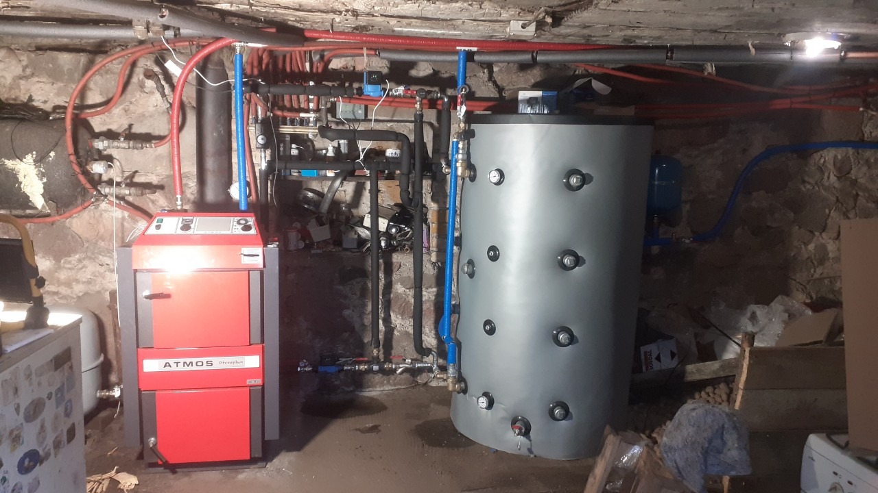 Czerwono-szary piec Atmos połączony z szarym zbiornikiem akumulacyjnym w piwnicy o kamiennych ścianach, z widocznymi rurami instalacji grzewczej i armaturą.