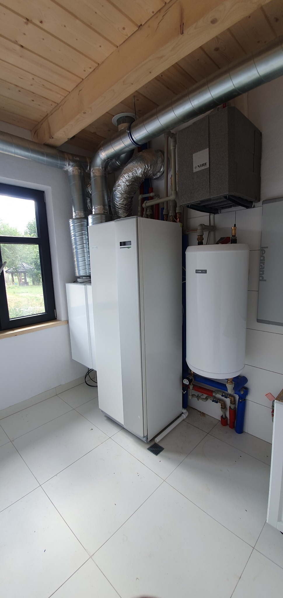 Biała jednostka centralna z szarym modułem wentylacyjnym NIBE, rurami i zbiornikiem na wodę w pomieszczeniu z drewnianym sufitem i oknem z widokiem na zewnątrz.