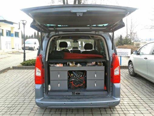 Wyposażenie warsztatowe w bagażniku samochodu typu kombivan: szuflady z narzędziami, organizer na drobne elementy, torba narzędziowa. Widoczny otwarty bagażnik, fragment parkingu z innymi pojazdami.