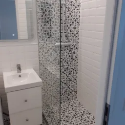 Biała umywalka z szafką podwieszaną i lustro na ścianie z białych płytek, kabina prysznicowa z czarno-białymi wzorzystymi płytkami na ścianie i podłodze.