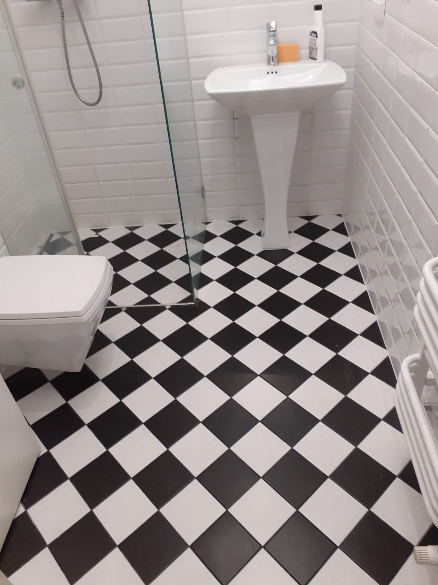Biała łazienka z czarno-białą podłogą w szachownicę, widoczna umywalka na postumencie, toaleta, grzejnik drabinkowy i kabina prysznicowa.