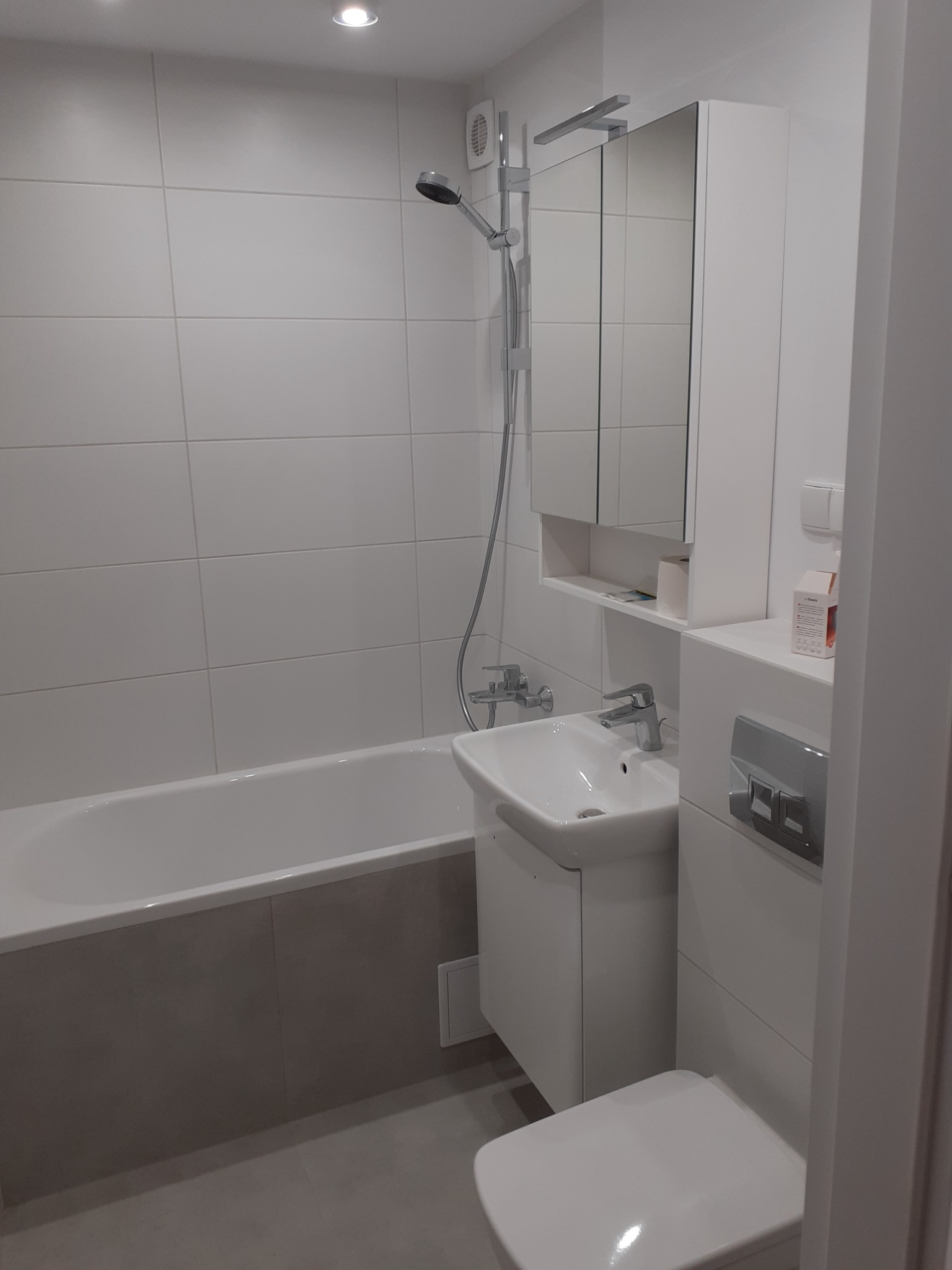 Nowoczesna łazienka z wanną obudowaną szarą płytką, białą umywalką z szafką, lustrem z oświetleniem i toaletą, wykończona białymi płytkami.