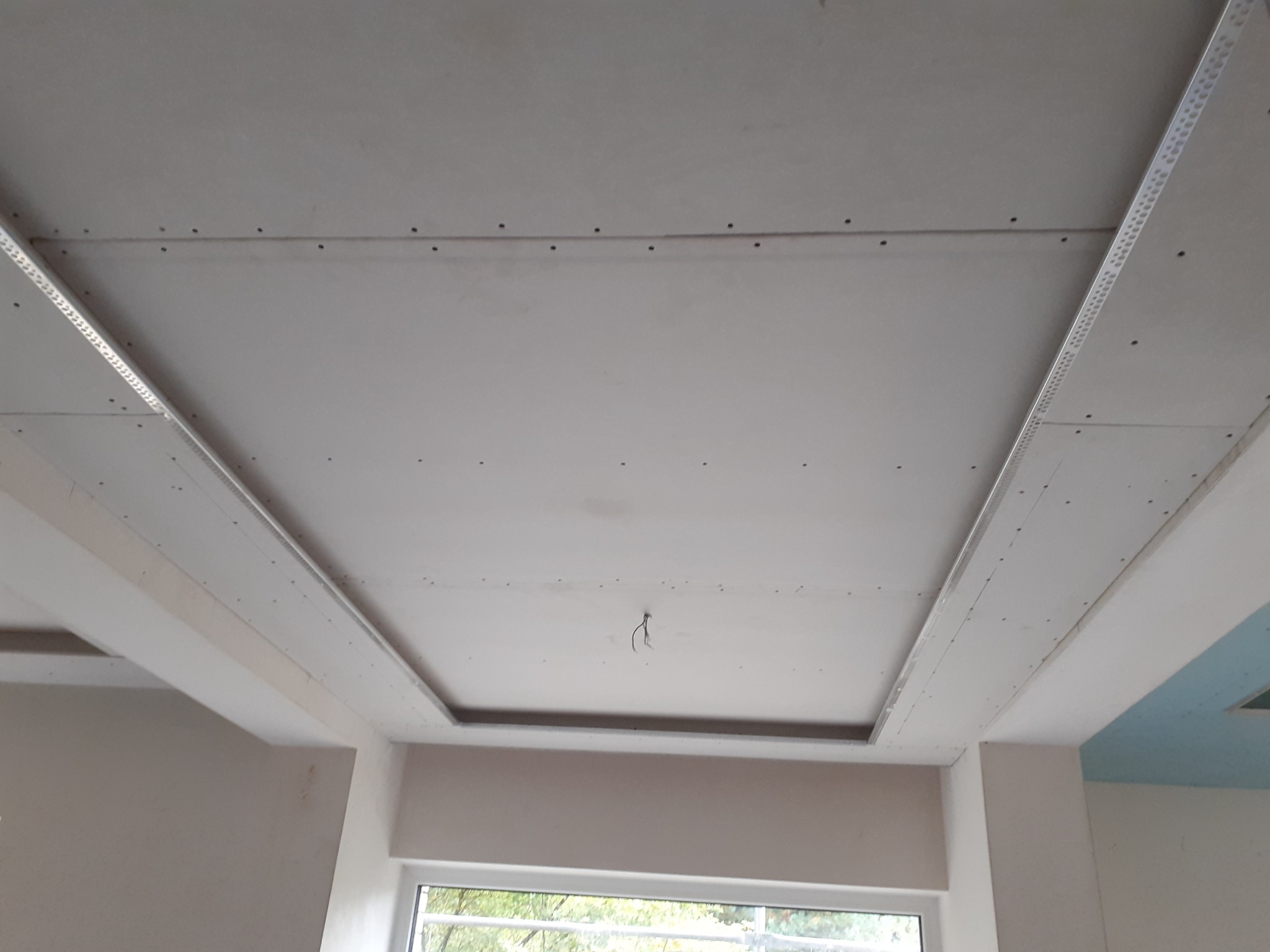 Montaż sufitu podwieszanego z płyty karton-gips z metalowymi profilami, widoczne ślady po wkrętach, fragment okna w tle.