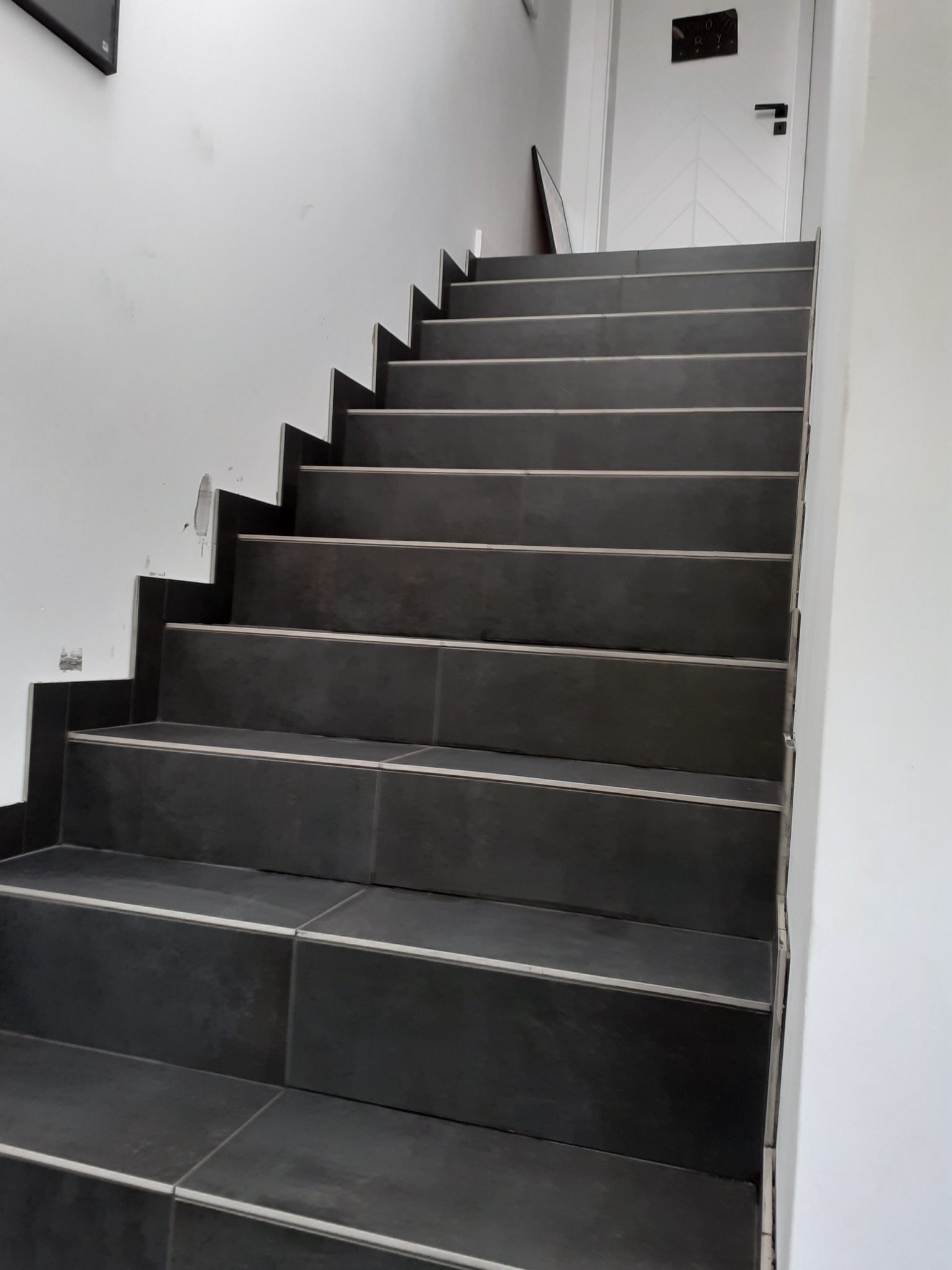 Ciemne, płytki ceramiczne na schodach z białymi listwami wykończeniowymi, widok z dołu na górę, biała ściana z widocznymi śladami po naprawach.