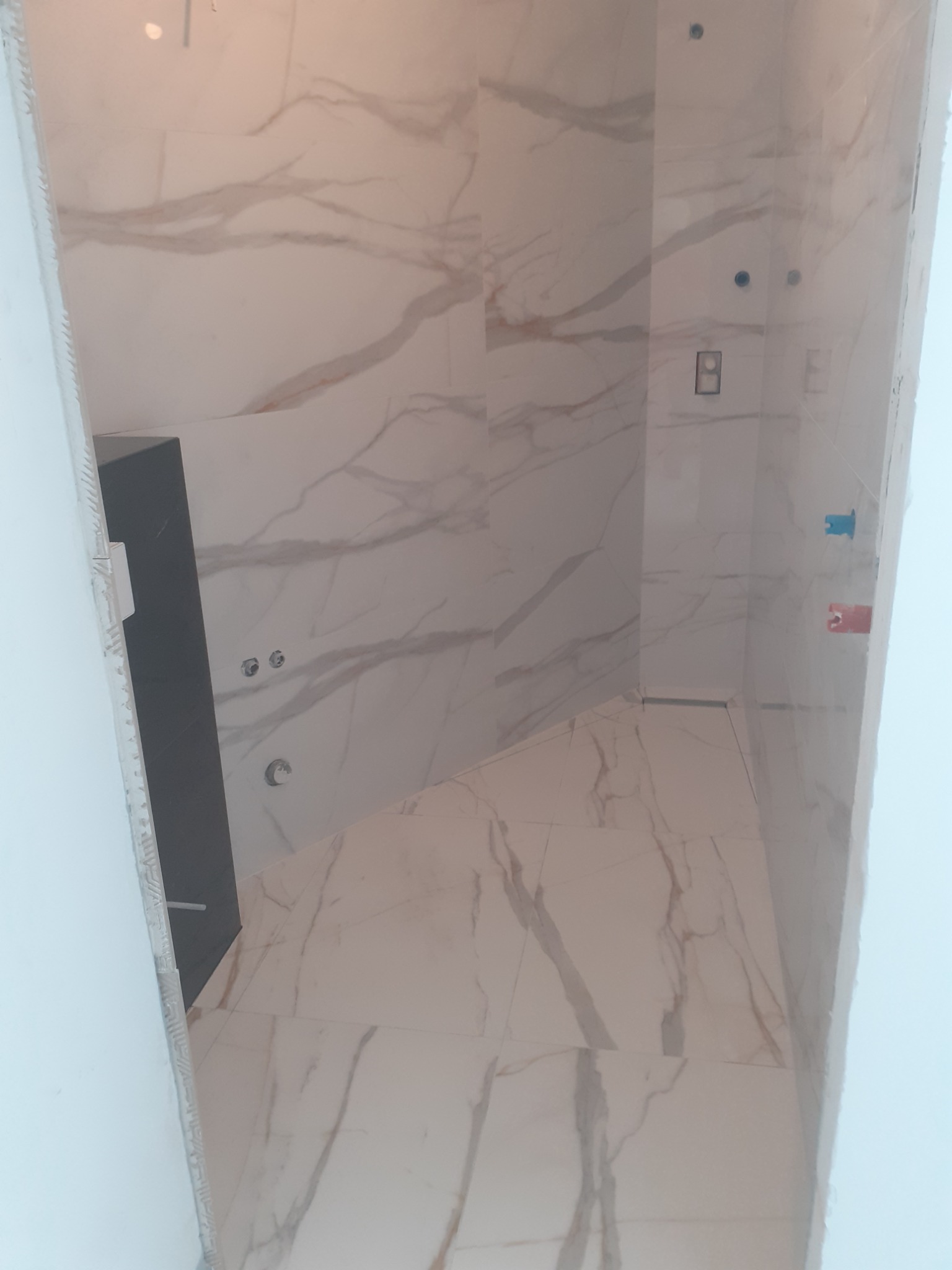 Łazienka w trakcie wykańczania, ściany i podłoga wyłożone płytkami imitującymi marmur, widoczne instalacje wodne i elektryczne, czarny element zabudowy.