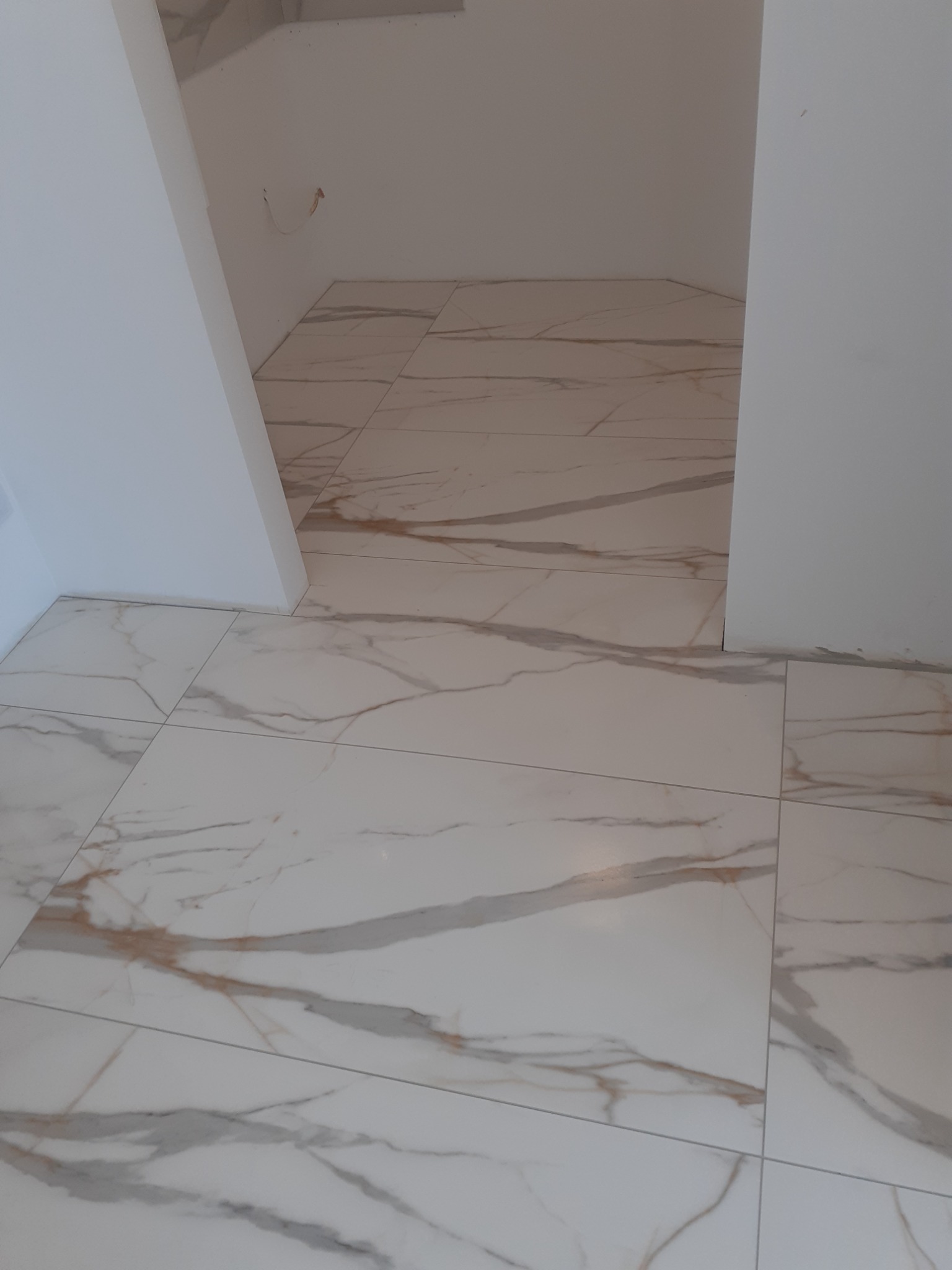 Ułożone, białe płytki podłogowe z szarymi i złotymi żyłami, imitujące marmur, w pomieszczeniu w trakcie remontu w Ostrołęce. Widoczne fragmenty ścian w kolorze białym.