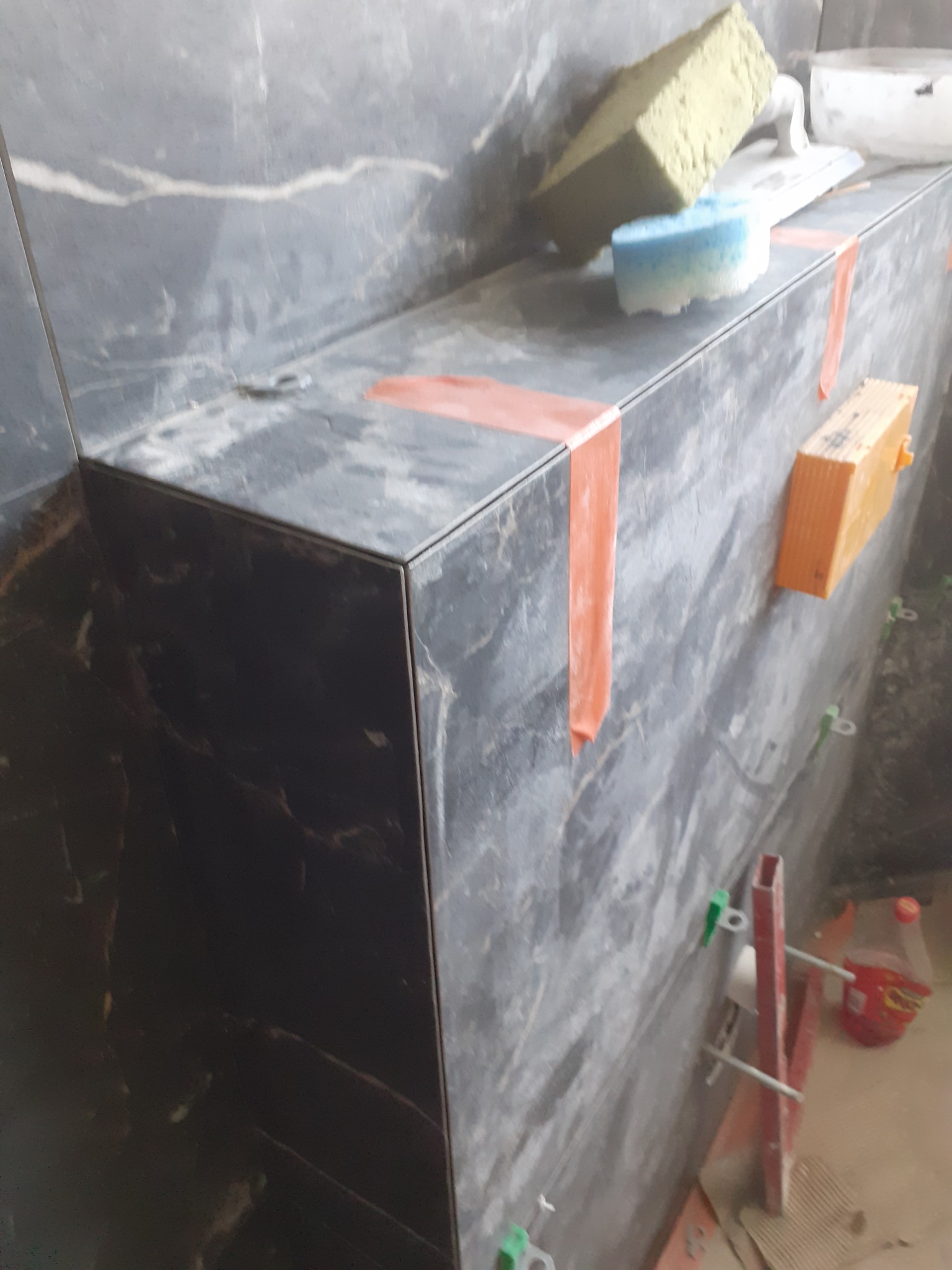 Ściana i półka wykończone czarnymi płytkami imitującymi marmur, z widocznymi narzędziami glazurniczymi: gąbką, szpachelką, taśmą malarską i systemem poziomowania płytek.