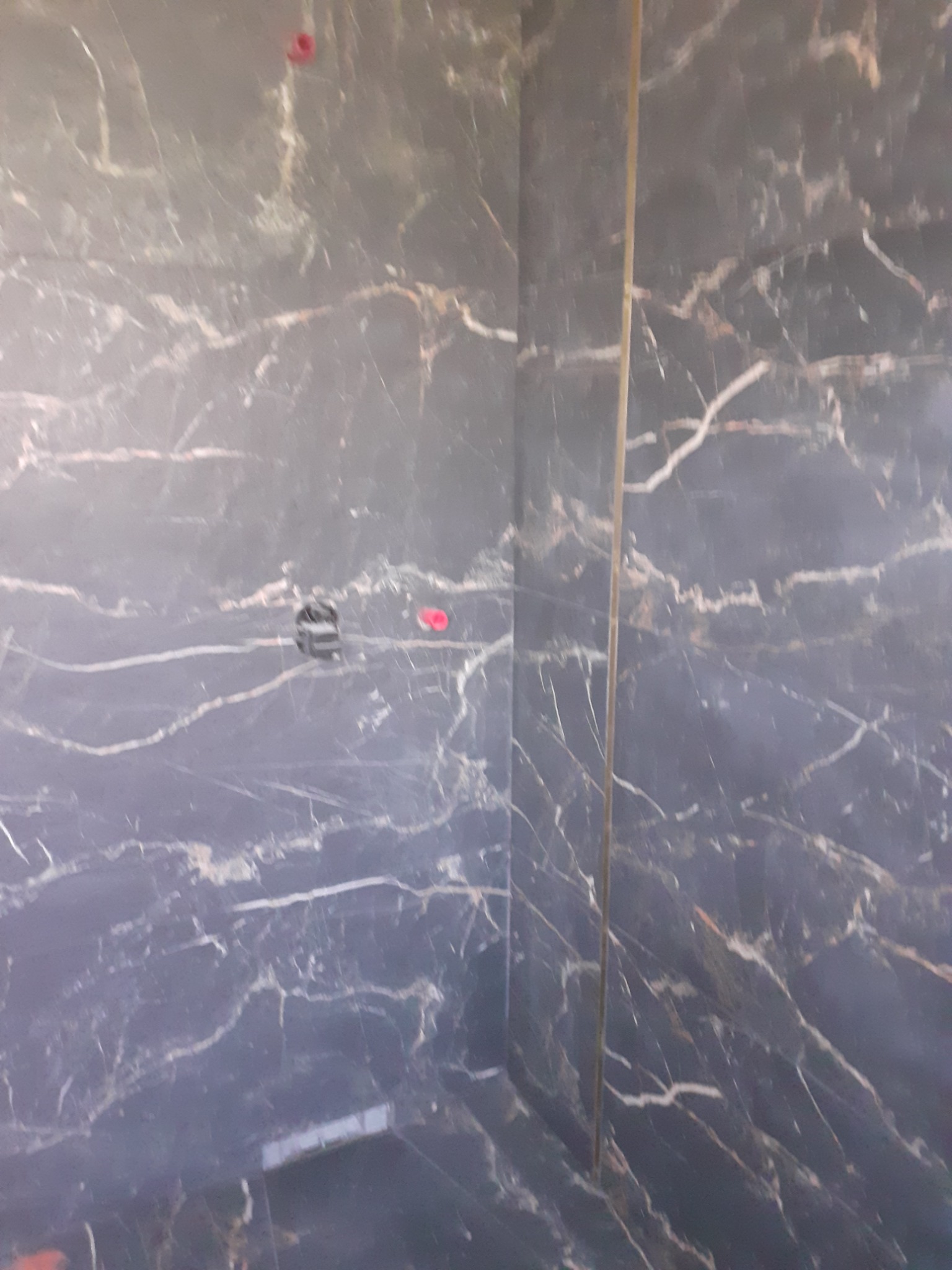 Ściana wyłożona ciemnymi płytkami imitującymi marmur z widocznymi białymi żyłkami, z fragmentem pionowego profilu wykończeniowego w kolorze złotym i elementami instalacji elektrycznej w ścianie.