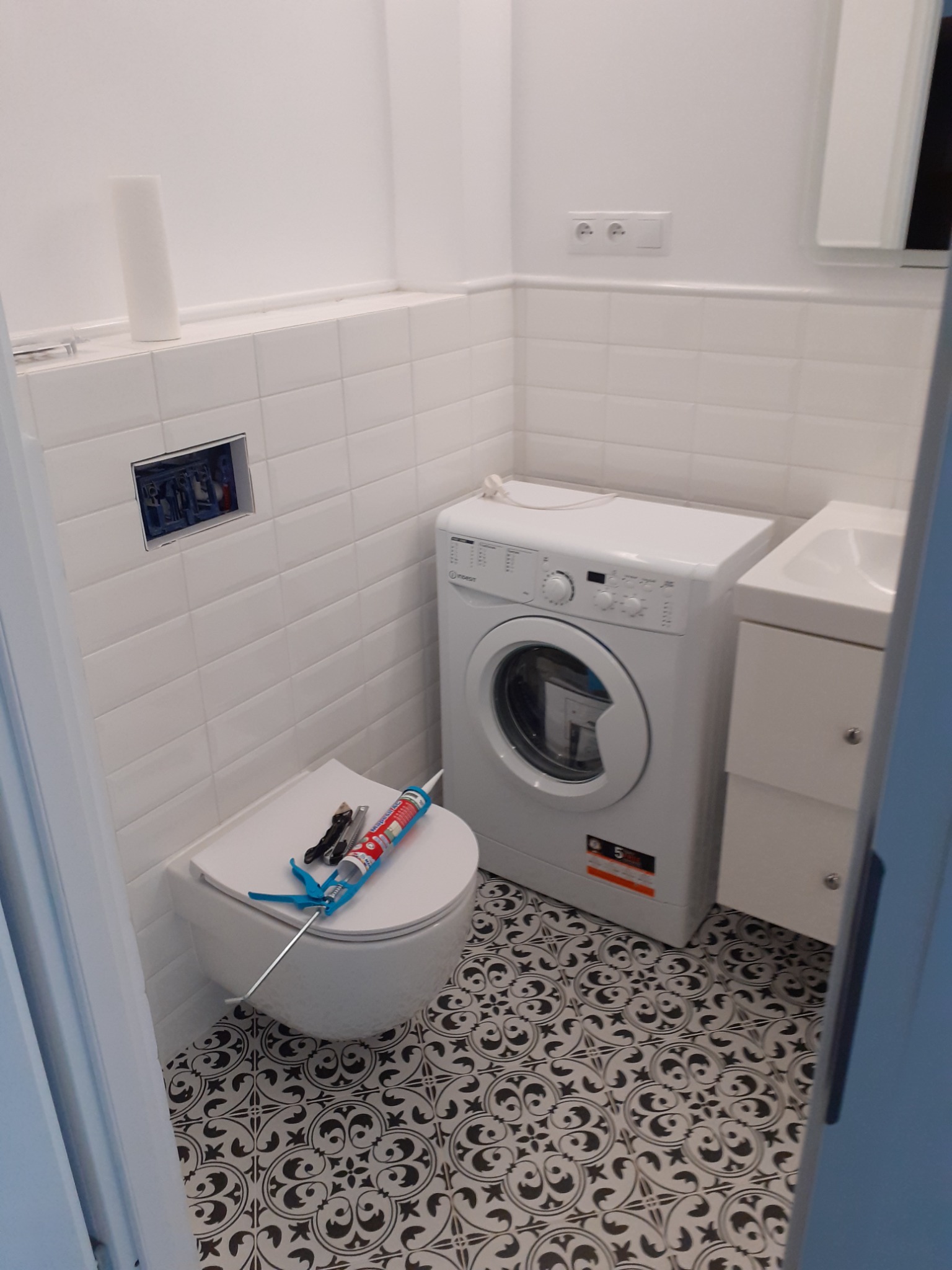Biała łazienka z umywalką, toaletą wiszącą i pralką. Na sedesie pistolet do silikonu i nóż. Podłoga w czarno-białe wzory.
