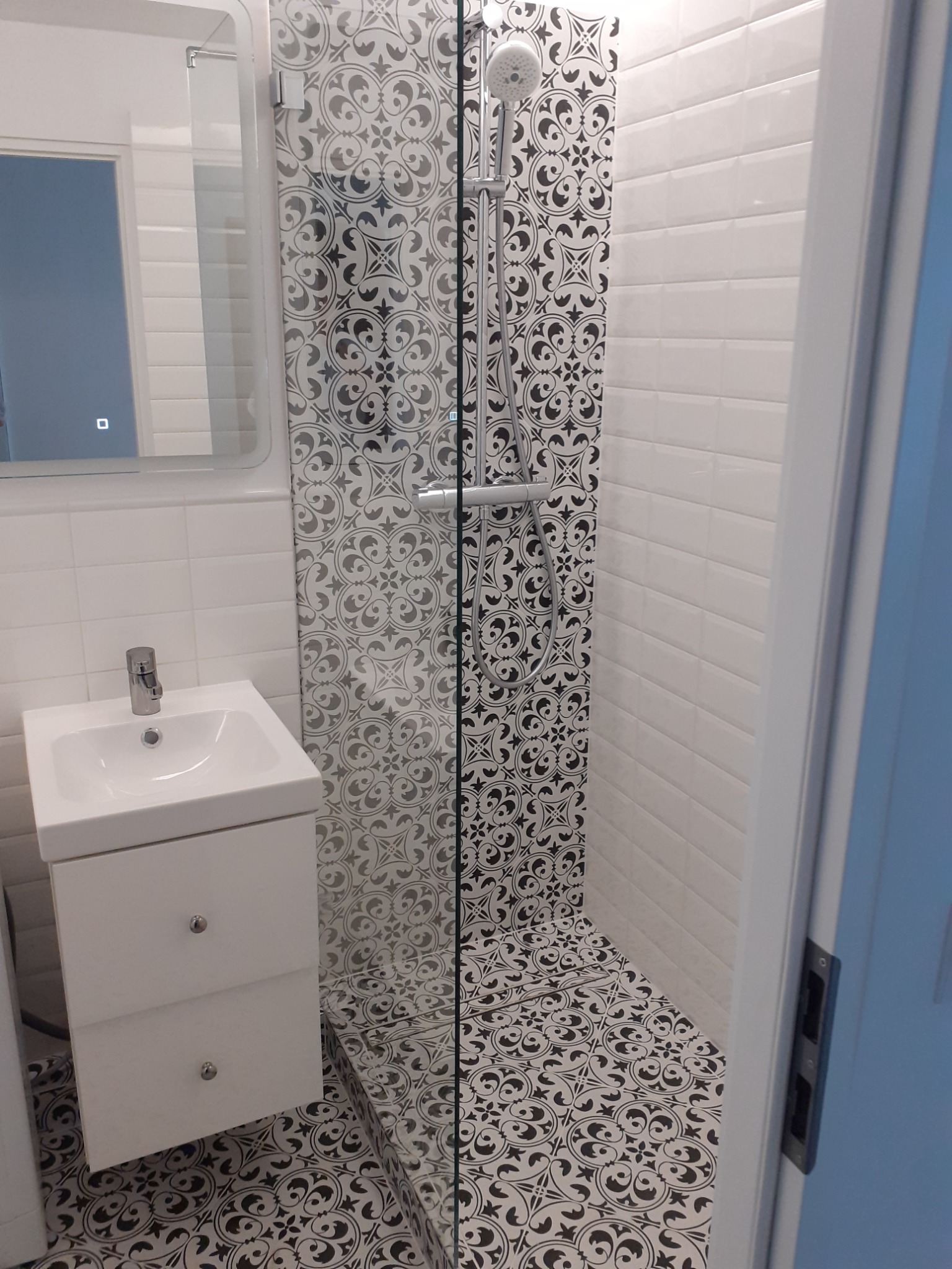 Biała umywalka z szafką podwieszaną i lustro na ścianie z białych płytek, kabina prysznicowa z czarno-białymi wzorzystymi płytkami na ścianie i podłodze.