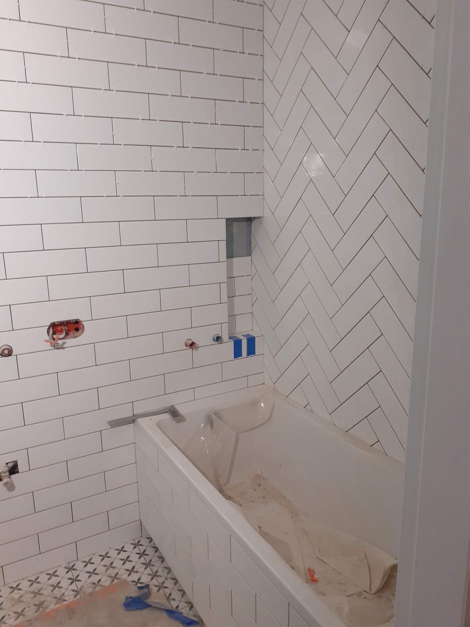 Białe, prostokątne płytki ułożone poziomo i w jodełkę na ścianach łazienki, widoczna wanna zabezpieczona folią, częściowo wykończona podłoga z wzorem w kwiatki, otwory pod armaturę.