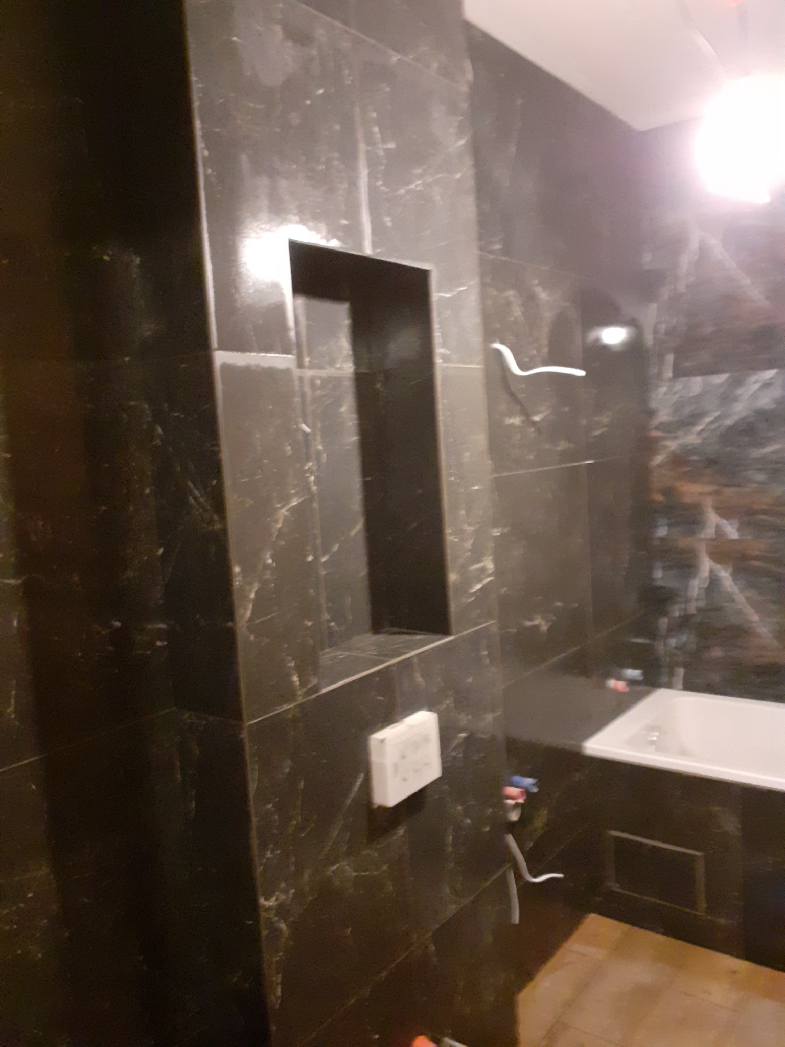 Ciemne, marmurkowe płytki w łazience z wnęką w ścianie, widoczny fragment wanny i biały włącznik.