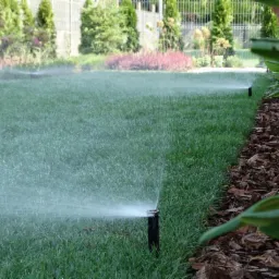 Automatyczny zraszacz nawadnia r&oacute;wnomiernie zielony trawnik, obok rabata wysypana korą.