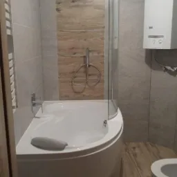 Nowoczesna łazienka z wanną narożną, szklanym parawanem, grzejnikiem drabinkowym i piecem gazowym na ścianie wyłożonej szarymi i drewnopodobnymi płytkami.